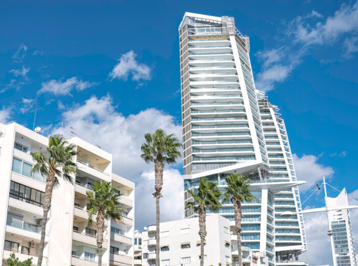 Modern skyscraper is being built, towering over traditional buildings with palm trees in Limassol, Cyprus